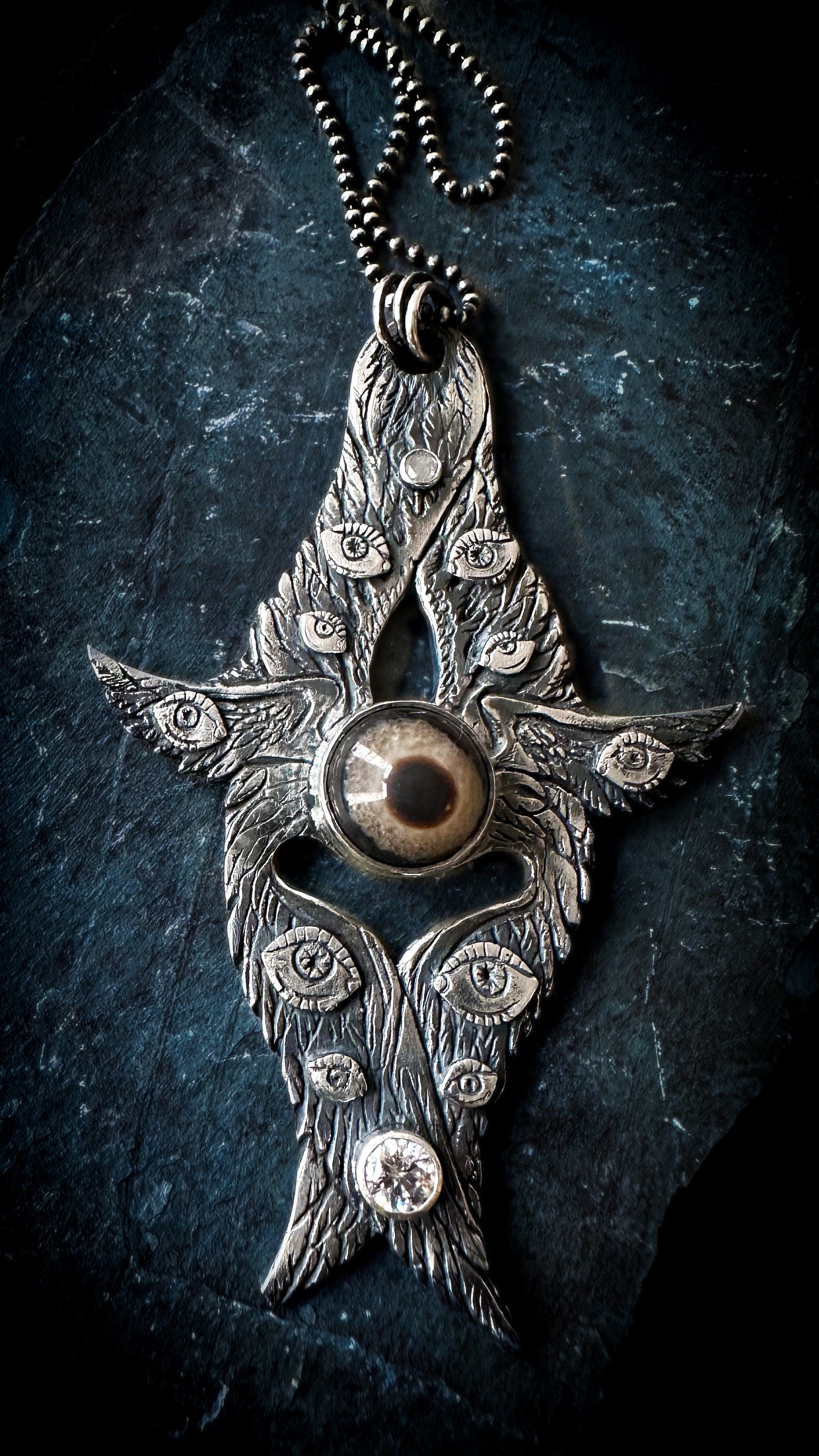 A medieval inspired, darkly gothic angel pendant in sterling silver. Fine craftsmanship with every feather distinctly carved. A realistic, natural agate ey, centrally bezel set and a salt and pepper diamond glitters at the top. Ten eyes and six wings, star and moons on the reverse side. By Silver Labyrinth Jewelry.