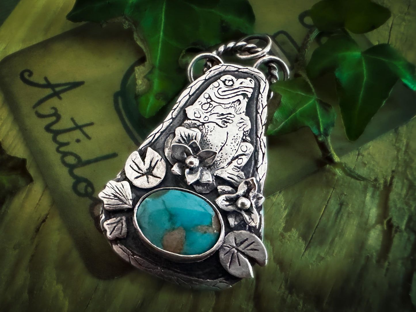 A little silver toad sits on a patinated backplate surrounds by flowers and lily pads. A gorgeous, all natural Carico Lake Mine turquoise is bezel set and sits beneath the toad. Handmade By Silver Labyrinth Jewelry