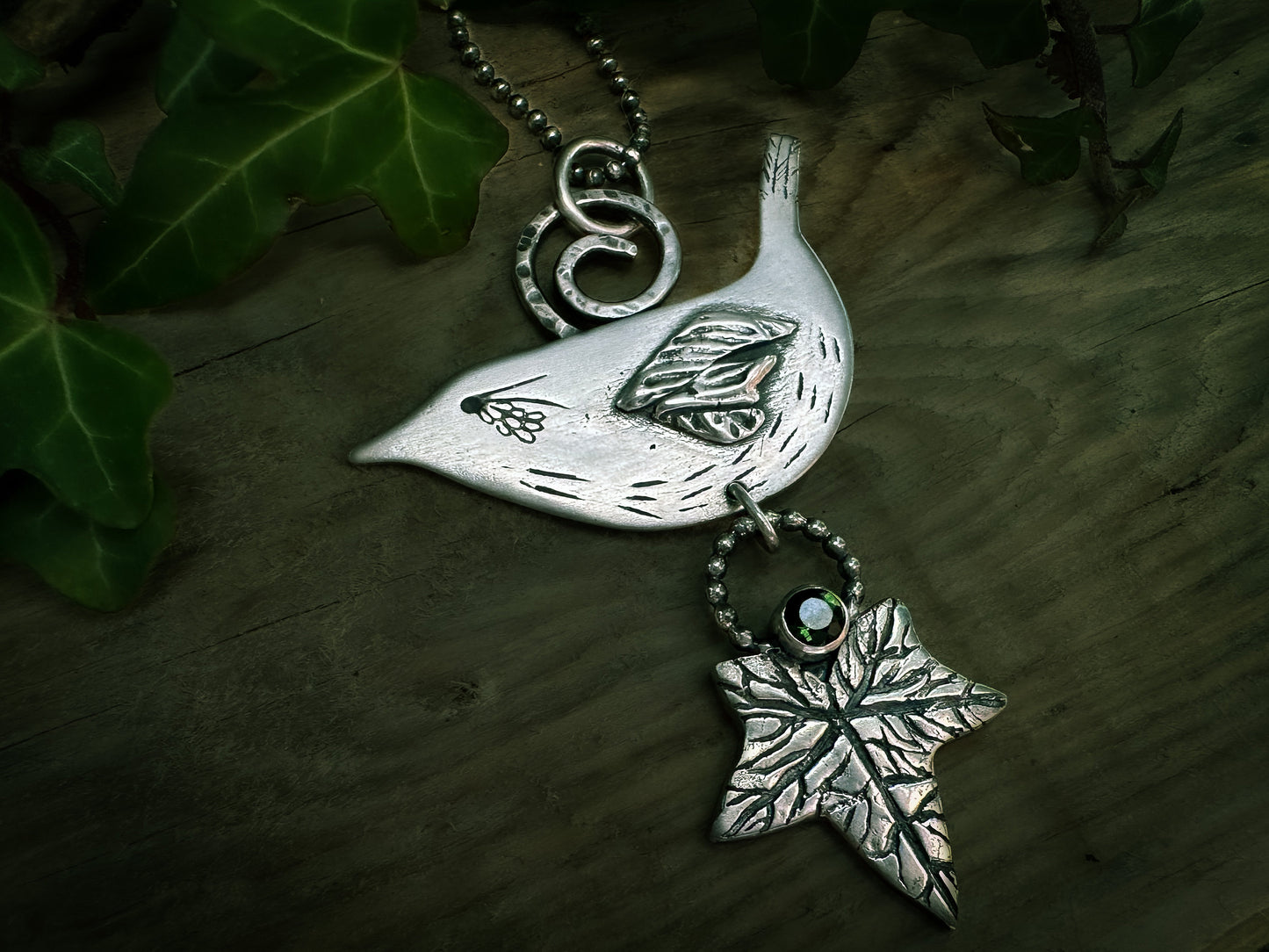 Elegant wren pendant, handmade in sterling silver and inspired by Celtic folklore. With detailed ivy leaf, hammered spiral bail and dark green tourmaline. By Silver Labyrinth