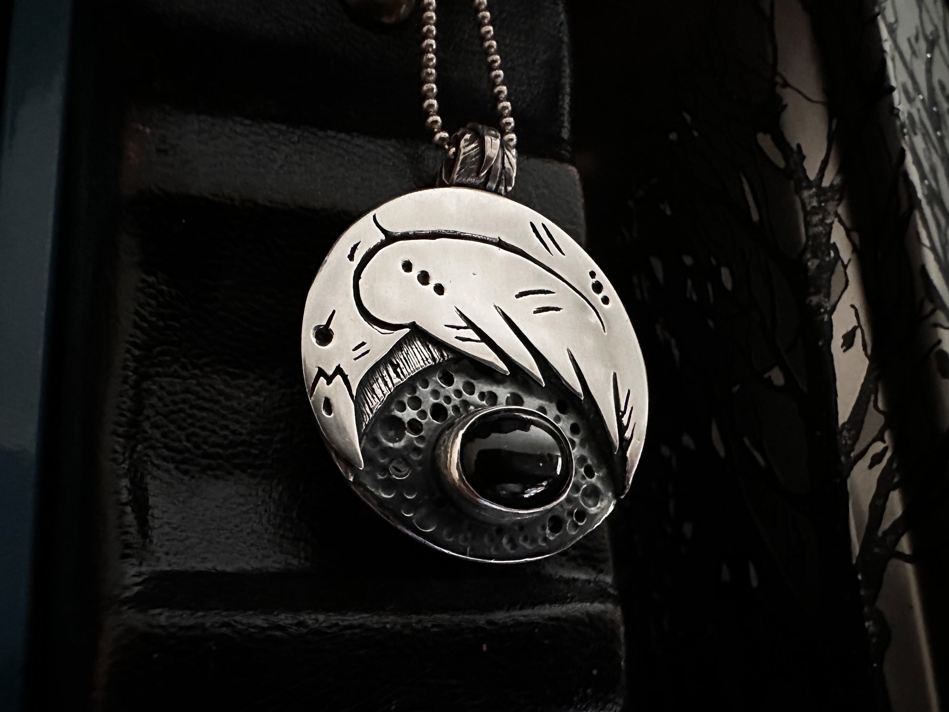 Oval silver crow pendant with textured background and a feather-detailed bail