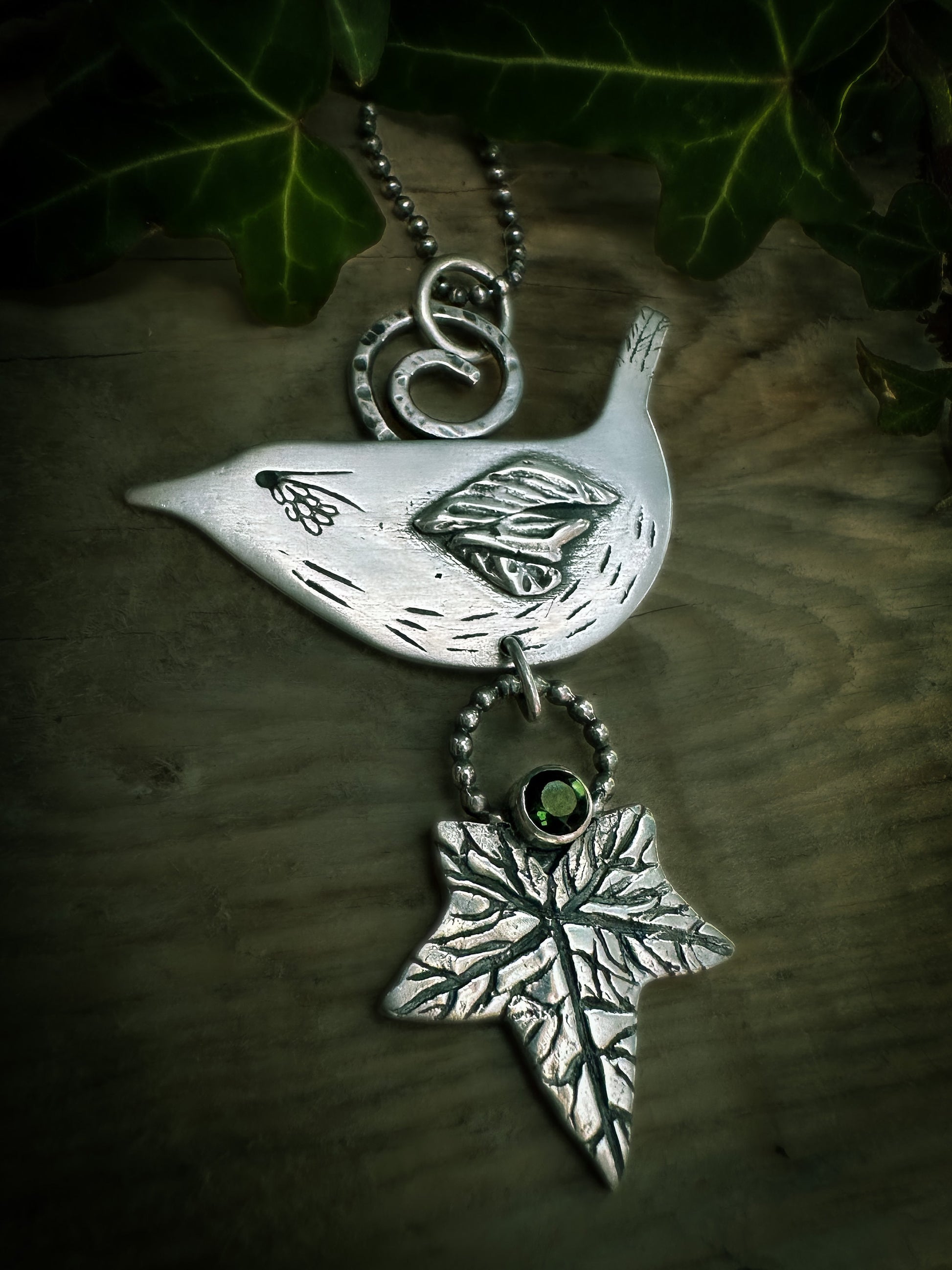 A detailed view of the sterling silver Druid Bird wren pendant. Finely detailed with a hammer spiral bail, realistic ivy leaf and a green tourmaline. By Silver Labyrinth Jewelry.