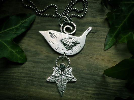 Druid Bird. A side-profile wren with detailed wing feathers sits on top of a detailed ivy leaf with a dark green, tube set faceted tourmaline. The back is decorated with spirals that reflect the ancient symbolism. By Silver Labyrinth Jewelry.