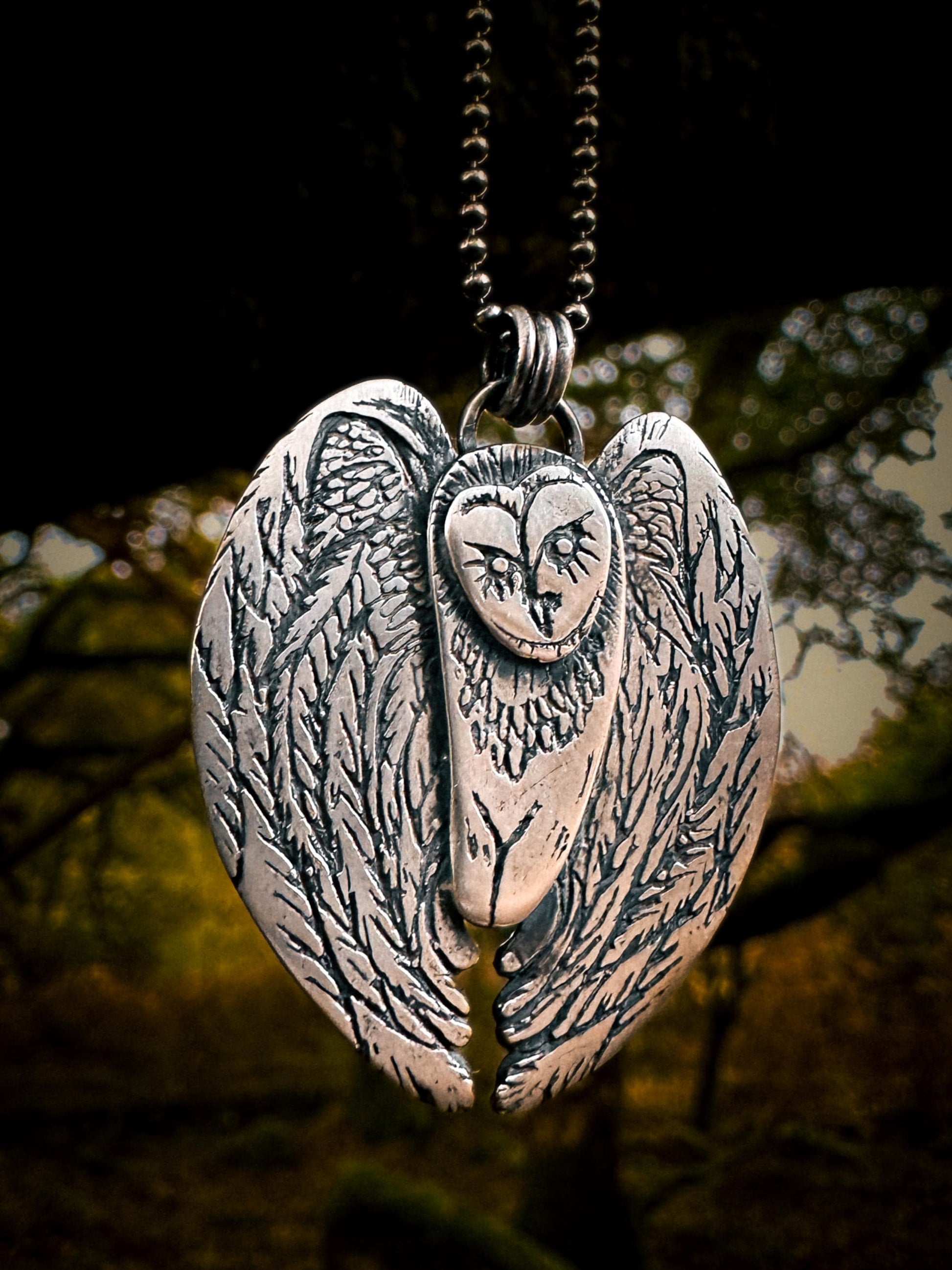 View of barn owl hanging from chain. The night owl has stylized features and an artisan feel. Its heart shaped face is full of expression ad each feather is detailed. Handmade in sterling silver 