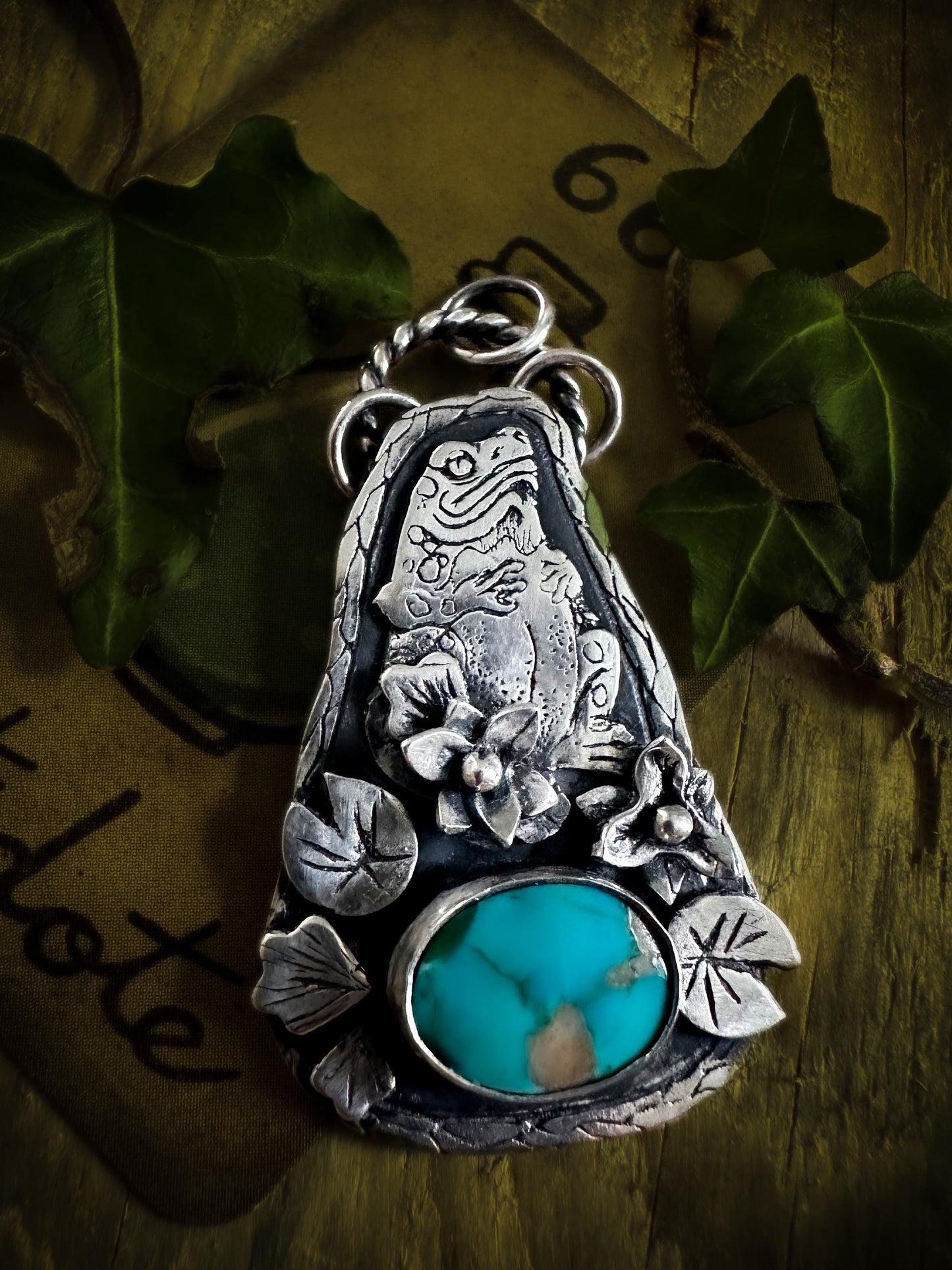 A sterling silver, handmade pendant of a toad sitting in a lily pond. The magical toad is surrounded two flowers and five leaves. It has been made with attention to details and set with a Carico Lake turquoise. Handmade by Silver Labyrinth Jewelry