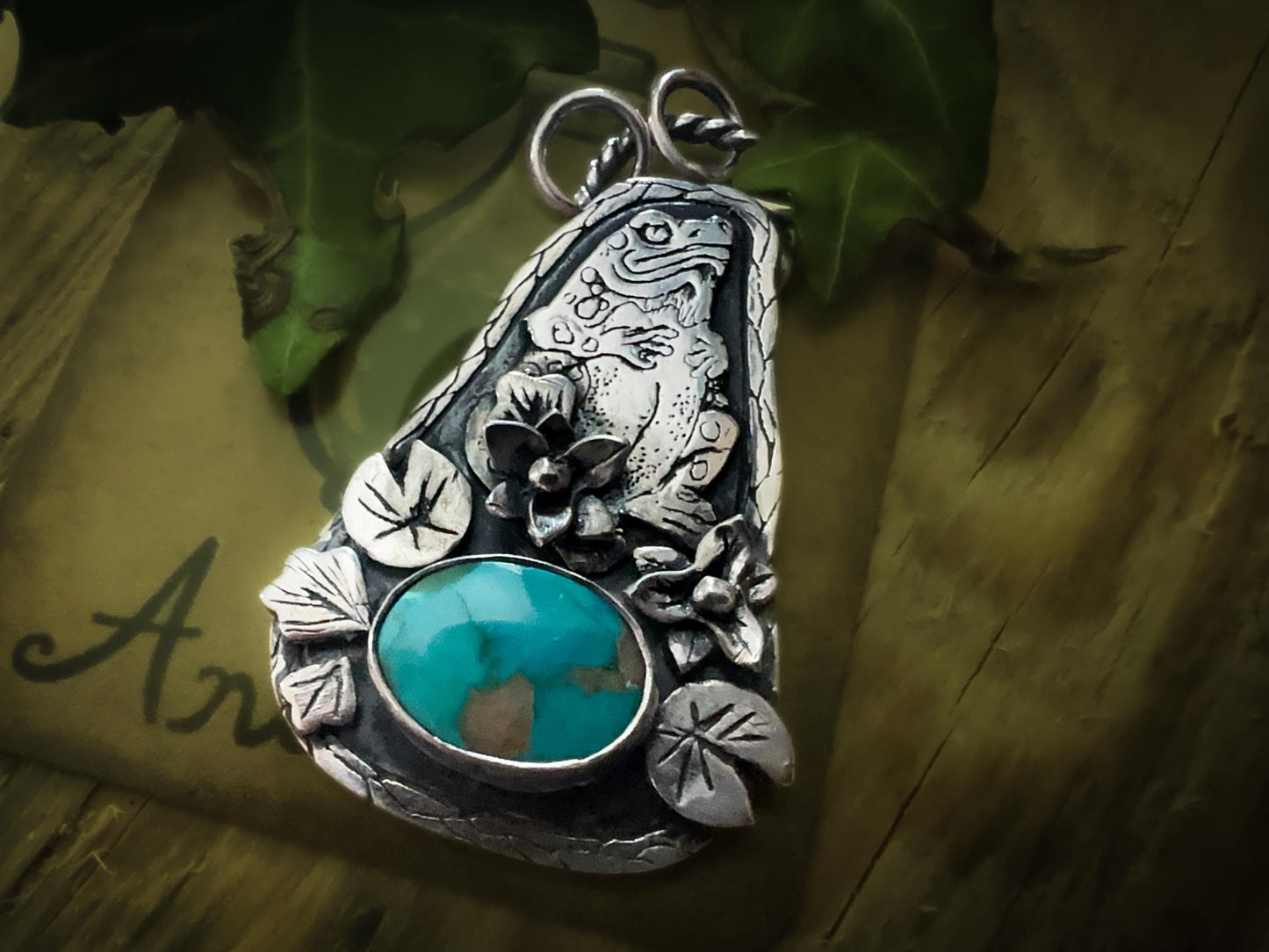 All natural Carico Lake gemstone sits beneath a toad totem animal. Surrounded by flowers and leaves. All handmade in silver. By Silver Labyrinth Jewelry