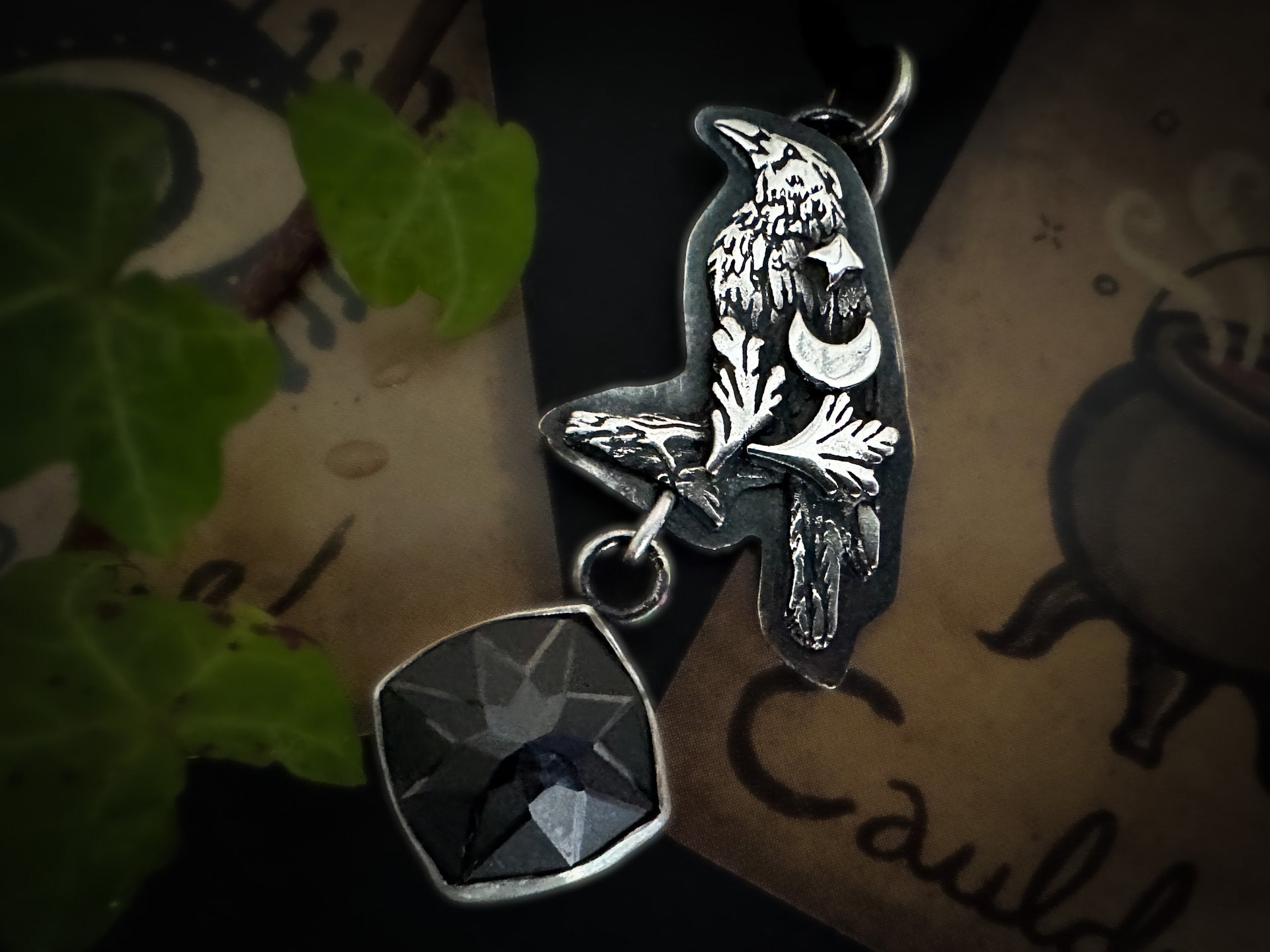 Silver crow necklace celebrating witches’ familiars, with crescent moon and star details and an engraved, faceted black spinel