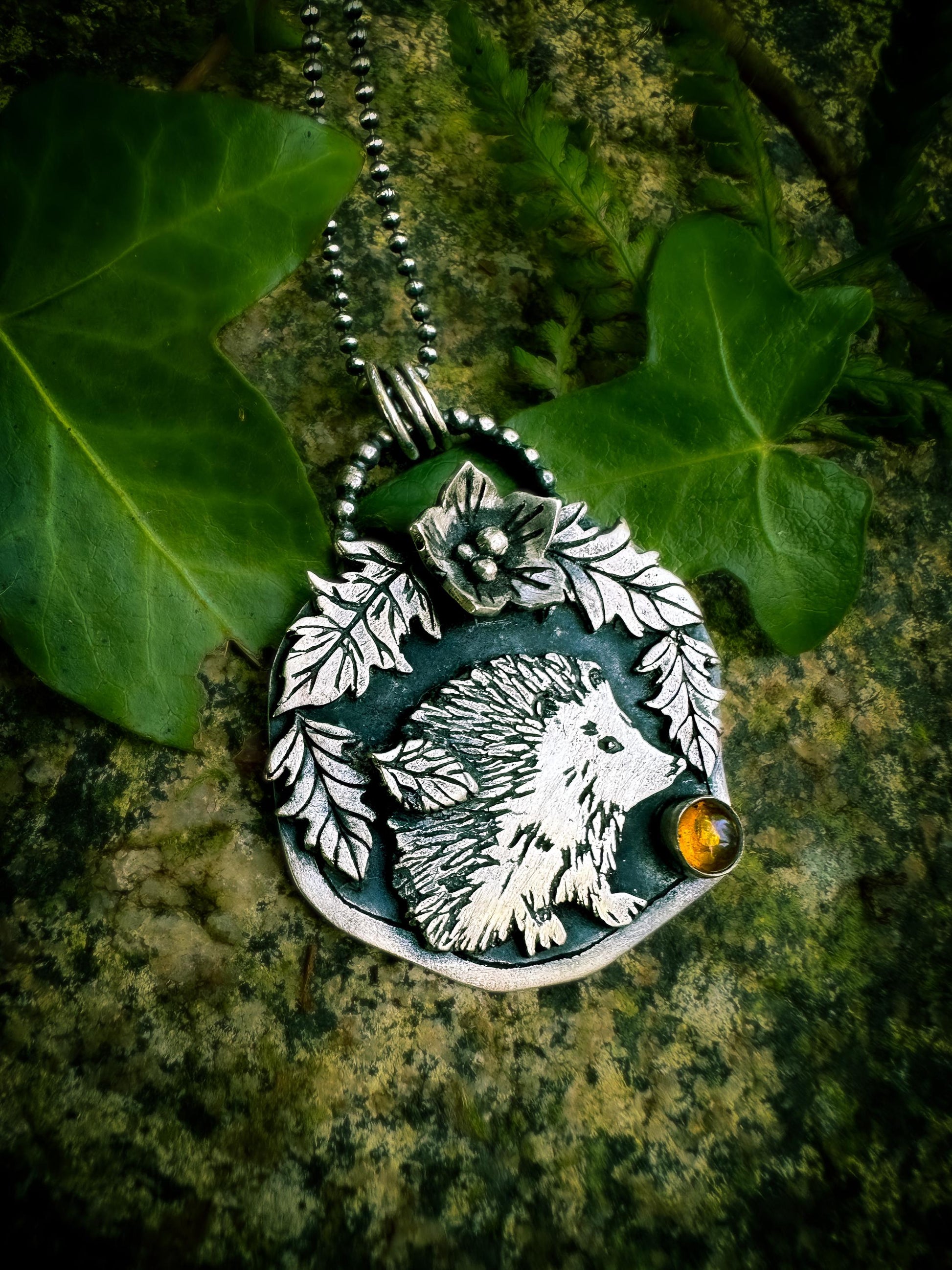 Side view of a little hedgehog with leaf details, a flower and amber handmade in sterling silver. 